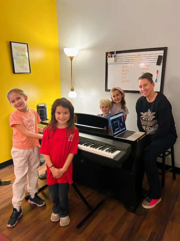 What are Group Instrument Classes at FAME Performing Arts Teacher Jen and smiling young students posing near a keyboard in a group instrument music lesson at FAME Performing Arts.