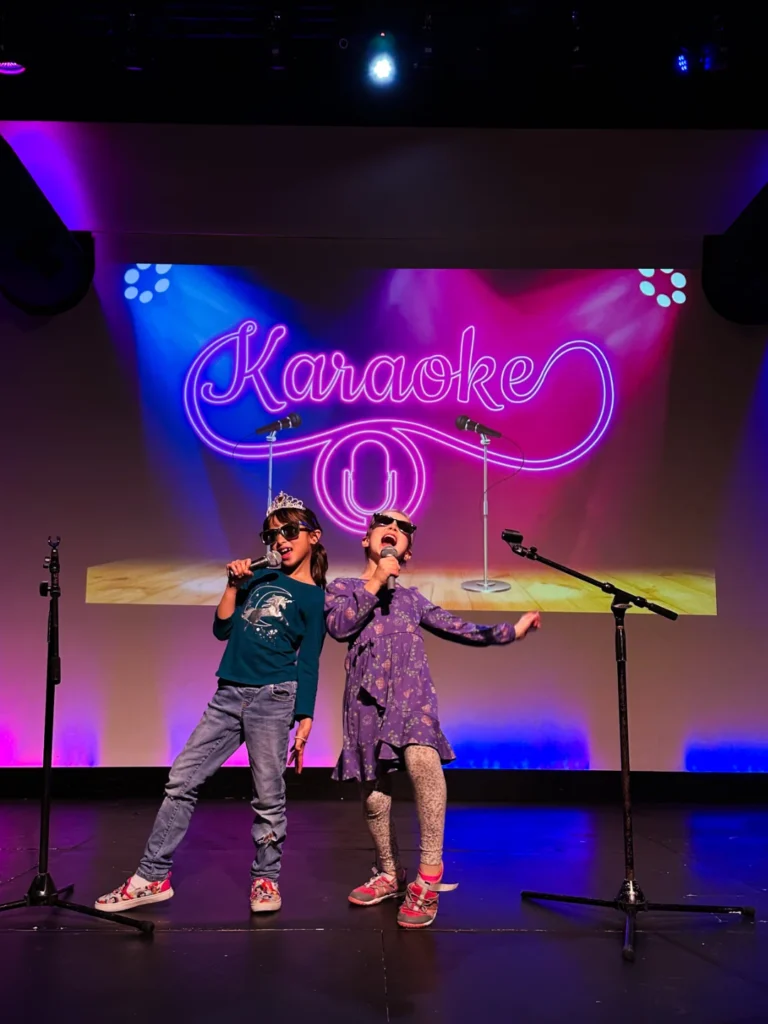 Three young girls in pink dresses singing karaoke on stage during a birthday party at FAME Performing Arts in Mount Pleasant.