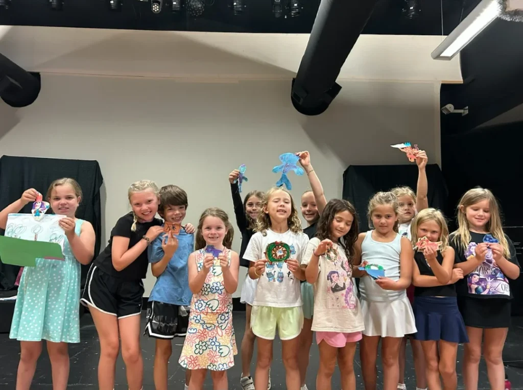 Group of summer camp students standing together on stage proudly displaying hand-crafted artwork.