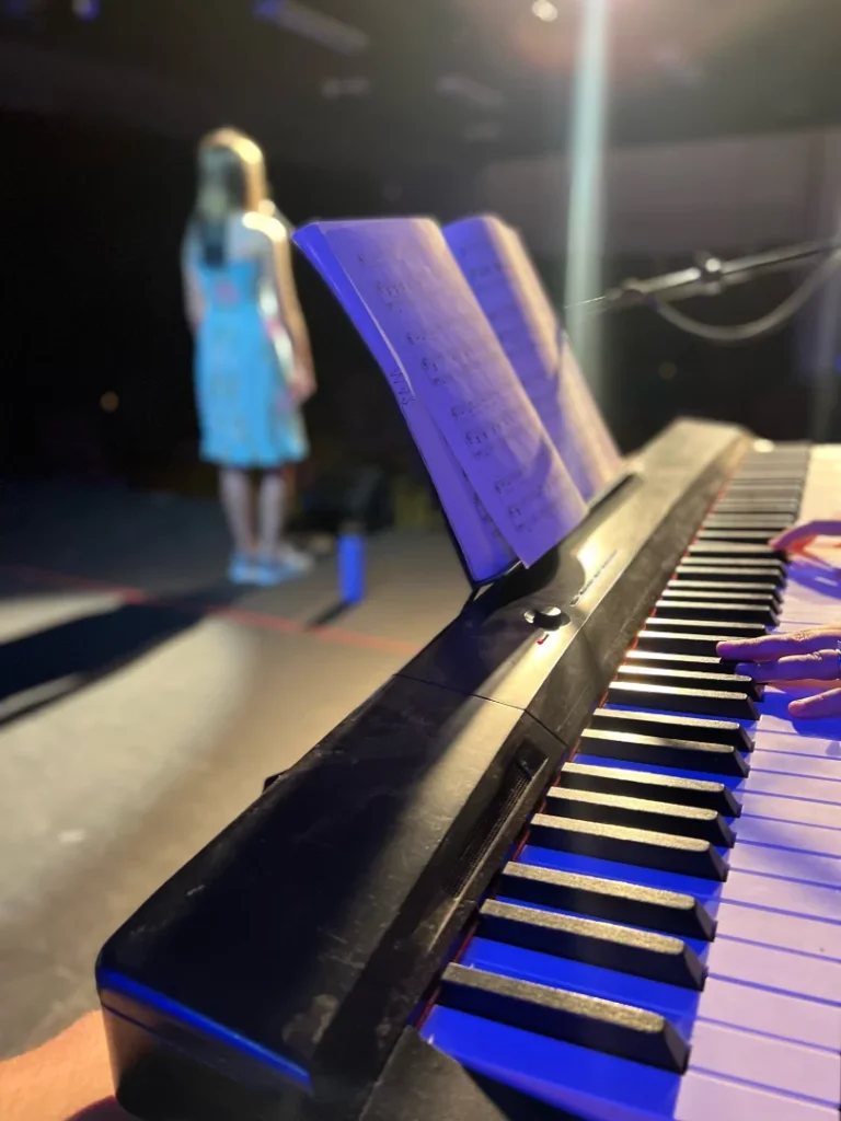 FAME - What Happens in a Voice Lesson Close-up view of piano keys and sheet music as an instructor accompanies a student singing on stage under warm lighting.